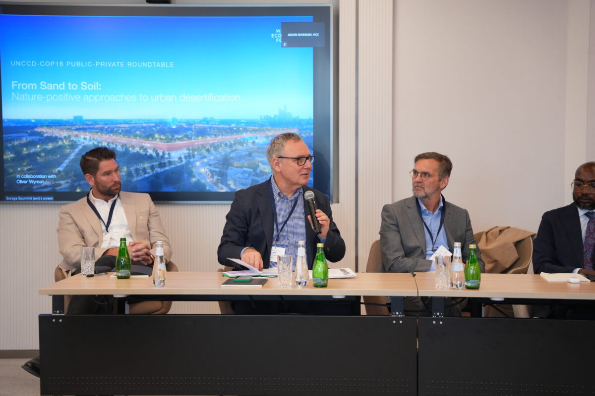 a man speaking at a microphone sitting at a table with other men